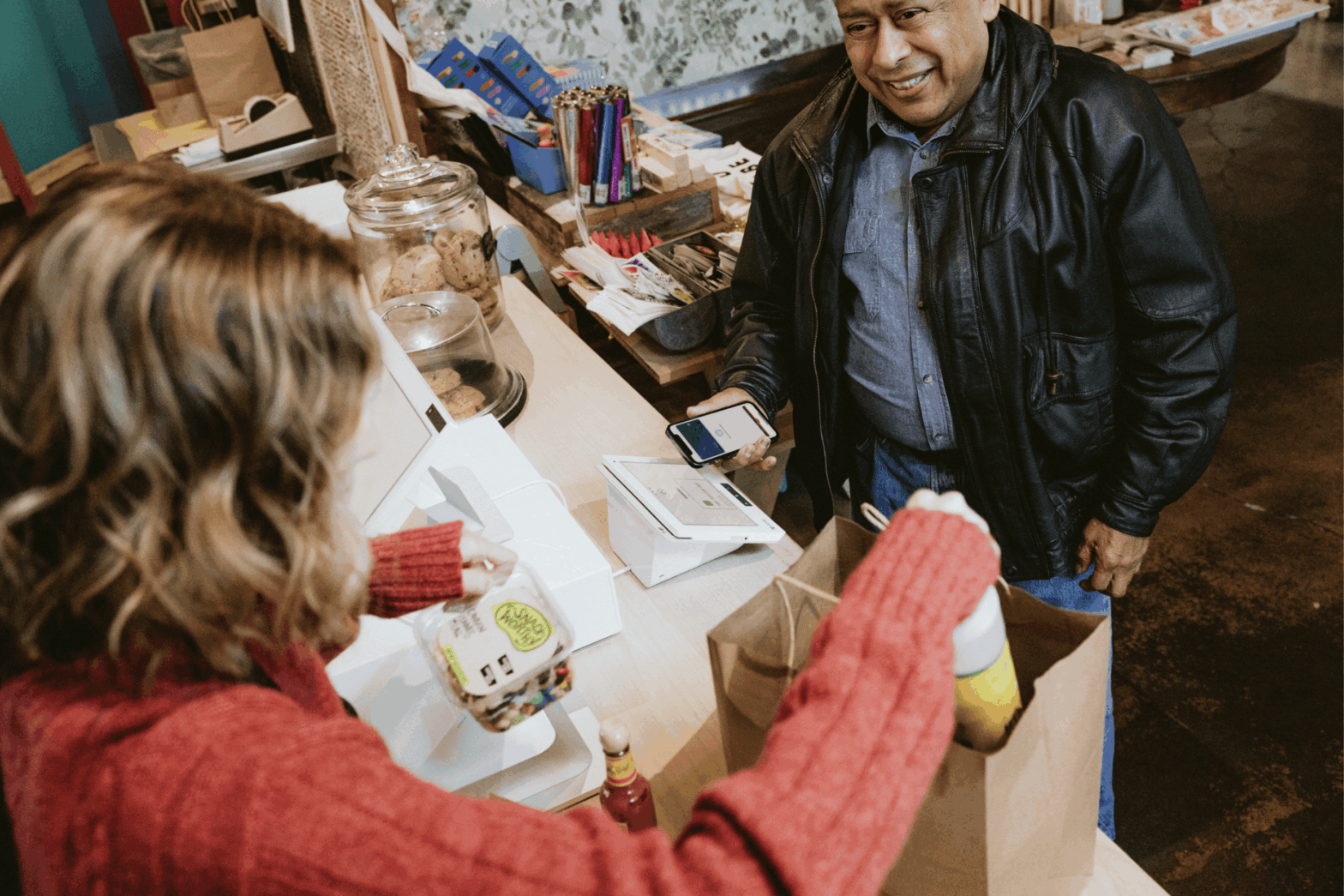 Smiling customer completing a contactless payment on a Clover Station Duo at a small retail shop counter, while the shop owner bags his order of cookies and snacks.